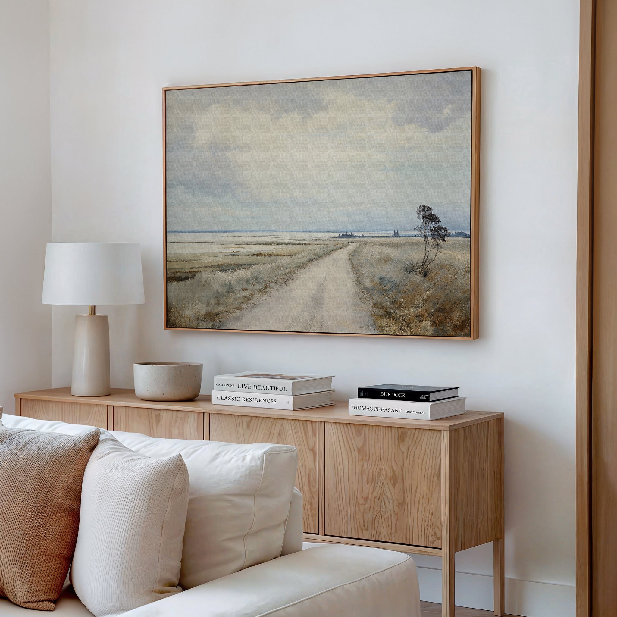 A cozy living room features a wooden sideboard with books, a lamp, and a bowl. Above it hangs the Far Light Canvas Art—minimalist wall art of a rural dirt road with fields and cloudy sky. A beige sofa sits in front.