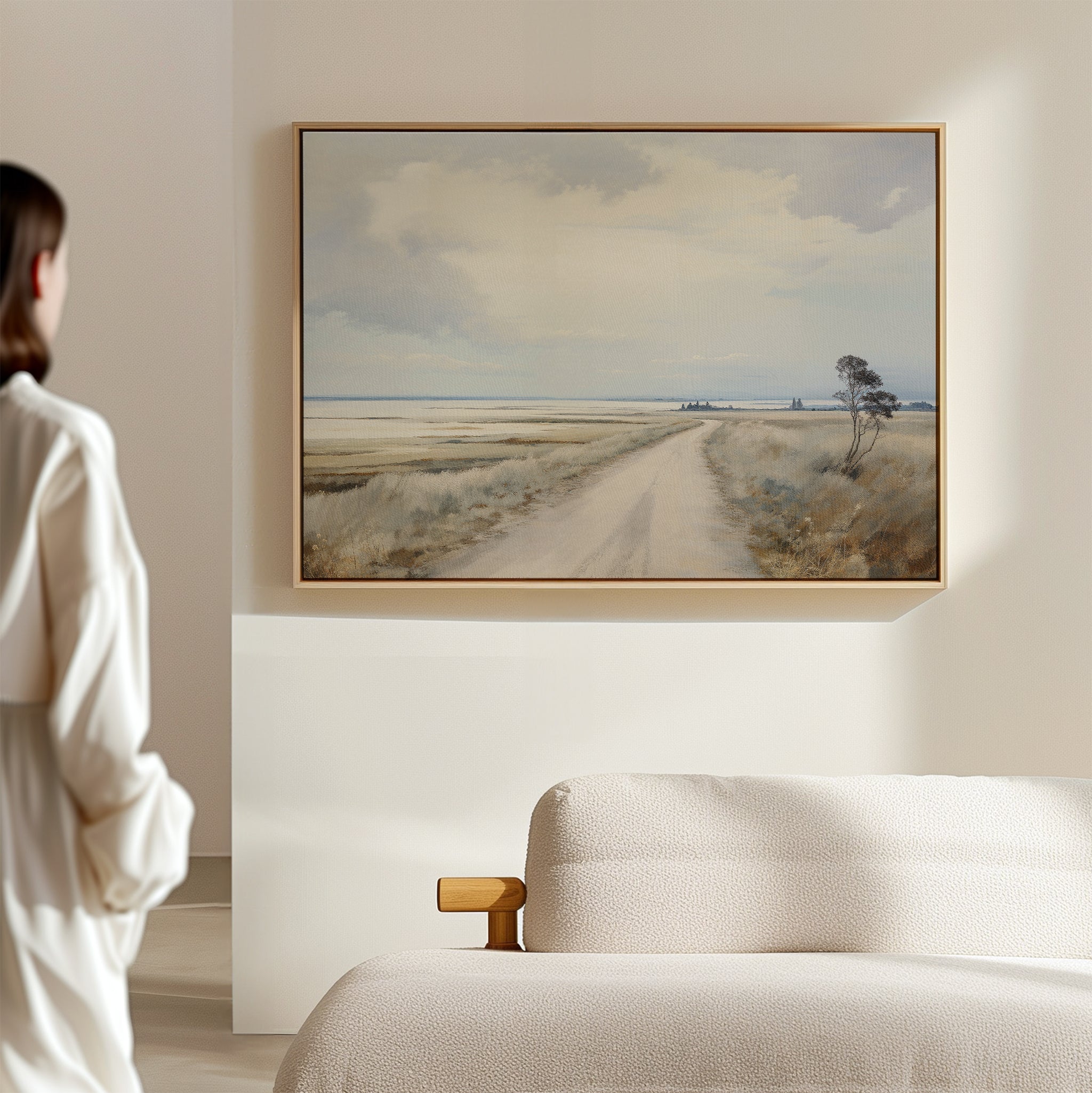 A person in a light outfit stands in a minimalistic room, gazing at the Far Light Canvas Art—a framed print of a dirt road through fields—hanging on a white wall above a beige sofa, embodying minimalist decor.