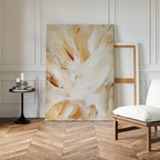 The Feathered Touch Abstract Canvas Art, featuring beige and white brushstrokes, leans against a white paneled wall. Nearby, an empty wooden frame, black table with candles, and cushioned chair enhance the elegant home décor.