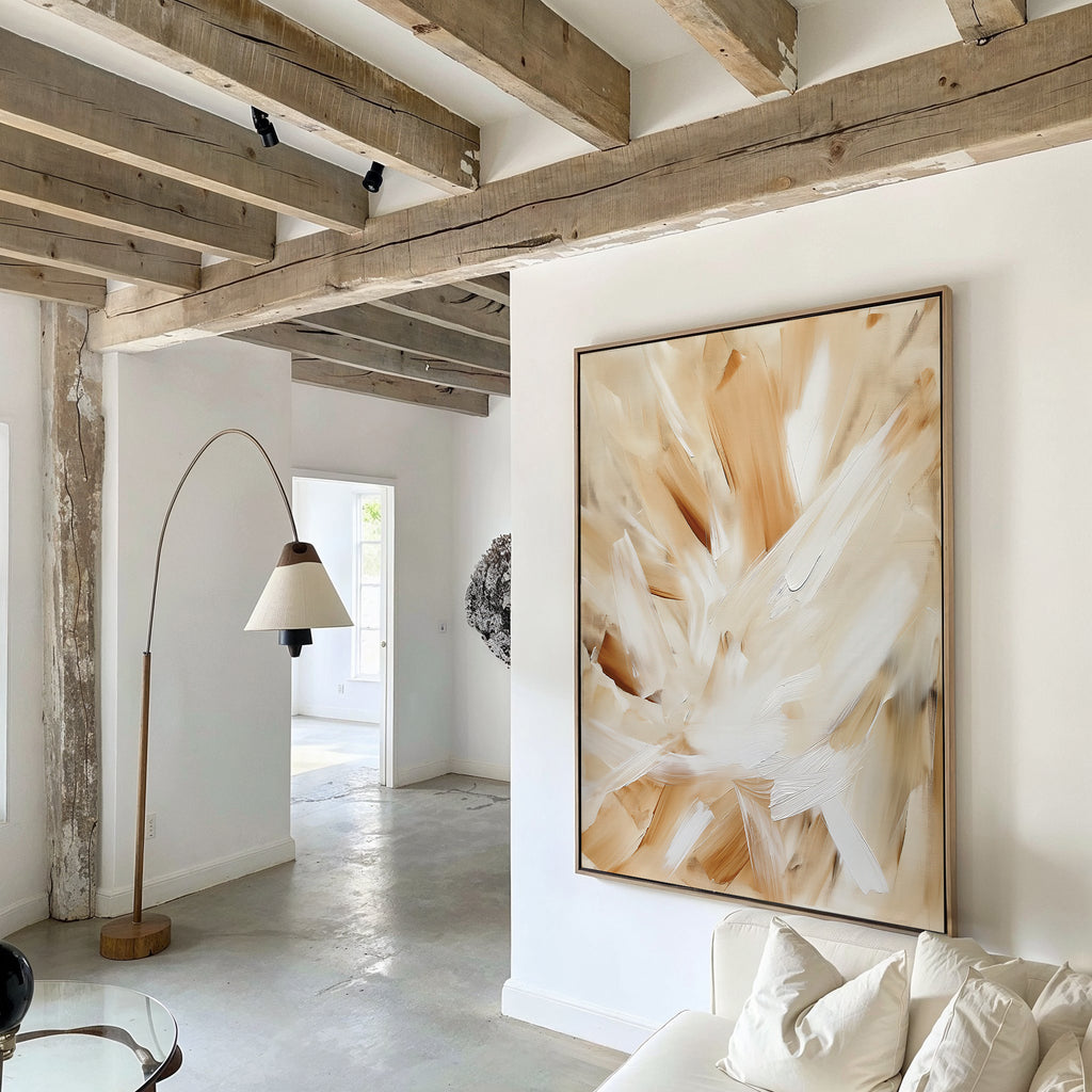 A modern living room features exposed wooden ceiling beams, elegant decor, the Feathered Touch Abstract Canvas Art in beige and white, a fabric-shaded floor lamp, white walls, and a white couch in the foreground.