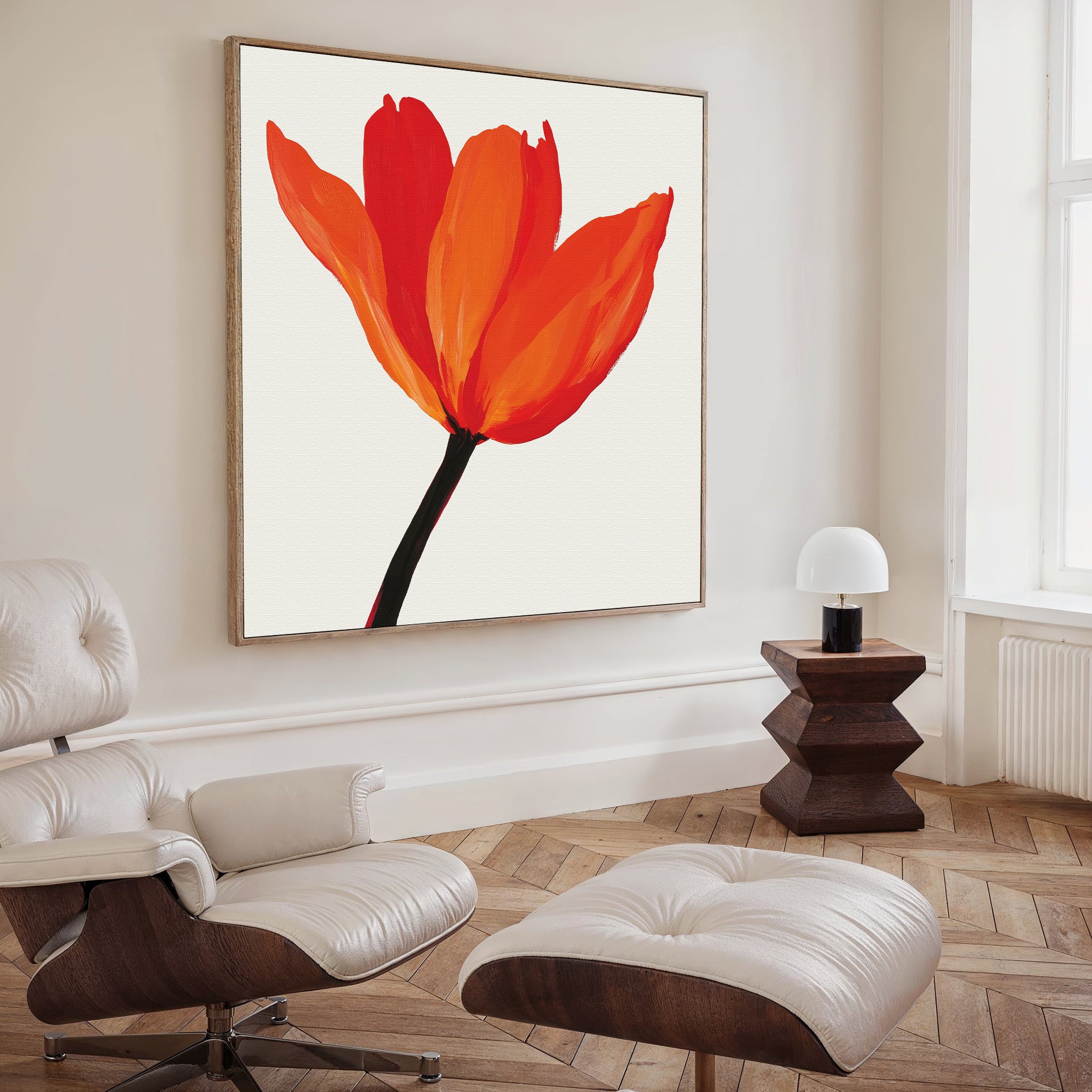 A modern living room features a white lounge chair and ottoman, dark wood side table with a white lamp, and striking Flame Tulip Canvas Art. Light wood floors and abundant natural light complete the minimalist decor.