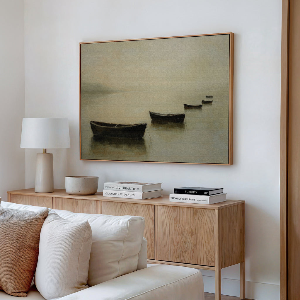 Minimalist living room with a light wood sideboard, books, white table lamp, bowl, and beige sofa. Serene decor is finished with the Floating Dreams Canvas Art of boats on calm water displayed above the cabinet.