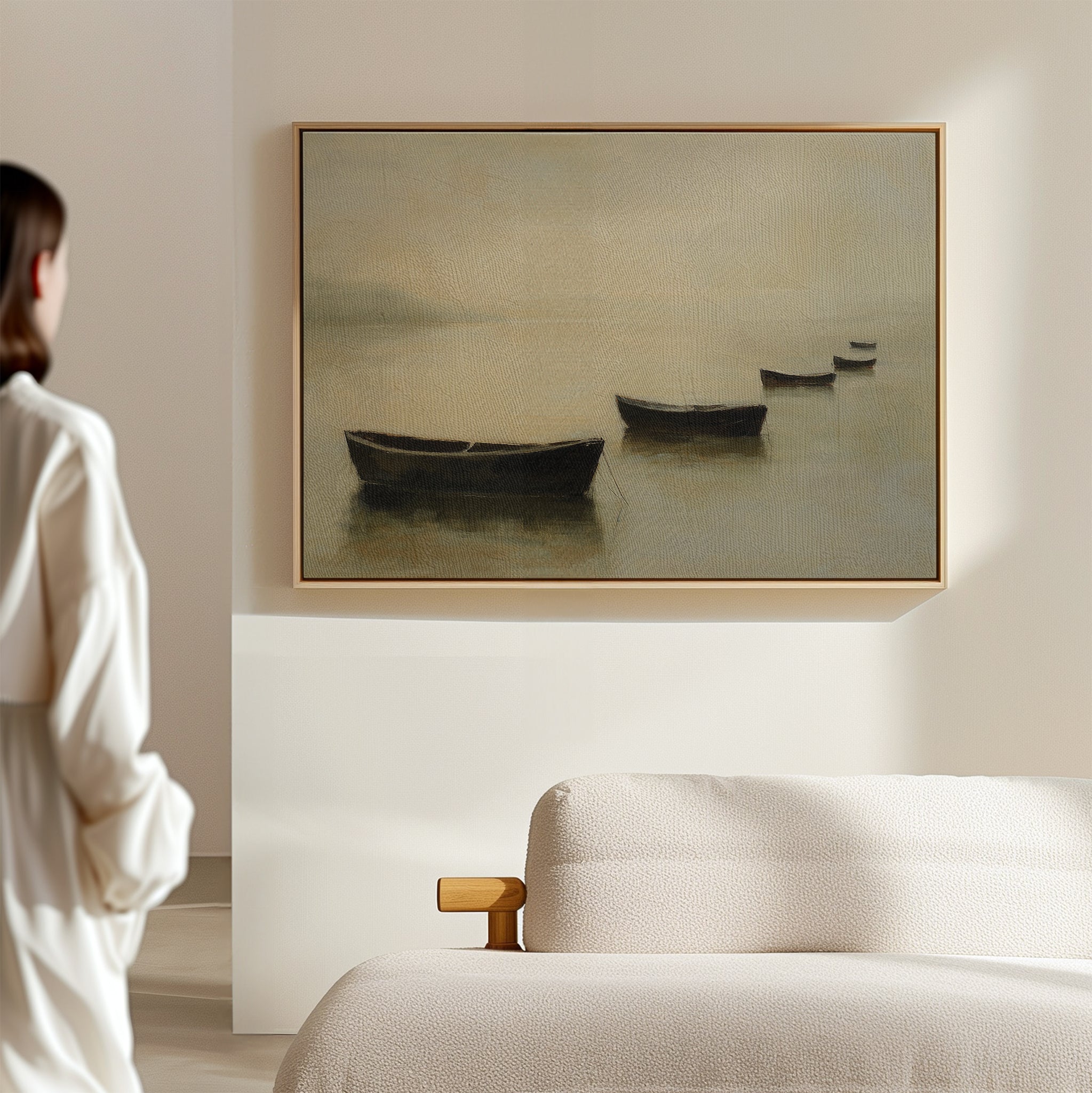 A woman stands in a minimalist room, gazing at the Floating Dreams Canvas Art—a painting of four rowboats on calm water—displayed above a beige upholstered sofa.