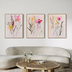 A modern living room featuring a curved beige sofa, round wooden coffee table, and the Floral Harmony Set of 3 prints in pink, yellow, and red on a white wall—perfect for chic living room decor.