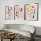 A modern living room featuring a curved light beige sofa, a round wooden coffee table, and the Floral Harmony Set of 3 vibrant floral framed paintings above the sofa—perfect for elevating your living room decor.