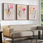 A modern living room features a beige sofa, round black coffee table, and the Floral Harmony Set of 3—framed floral wall art in pink, yellow, and green—adding chic style to the white walls.
