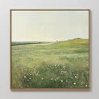 The Floral Meadow Canvas Art, featuring a sunlit grassy meadow with white wildflowers, rolling hills, and a distant tree beneath a pale sky, hangs on a white wall—an ideal touch for living room decor.