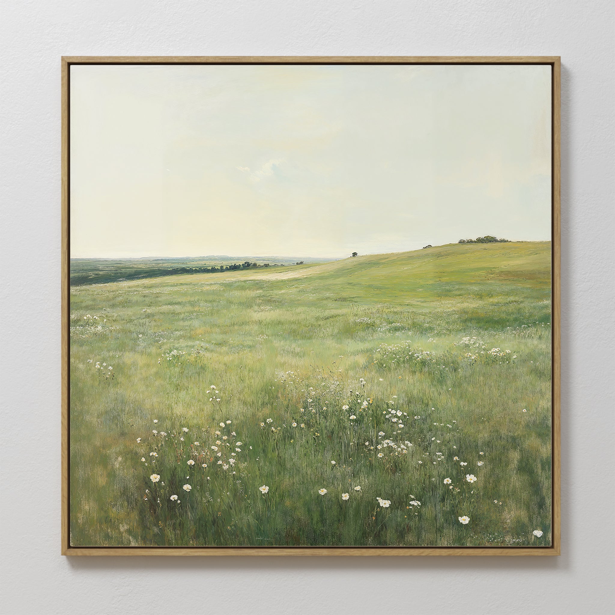 The Floral Meadow Canvas Art, featuring a sunlit grassy meadow with white wildflowers, rolling hills, and a distant tree beneath a pale sky, hangs on a white wall—an ideal touch for living room decor.