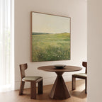 A minimal dining area features a round wooden table, two cushioned chairs, and the Floral Meadow Canvas Art on a white wall. Sunlight through sheer curtains creates a thoughtful living space for decor enthusiasts.