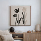 A minimalist living room features a wooden sideboard, neutral decor, and the Floral Simplicity Canvas Art—a framed print with three black and beige tulip-like flowers—adding elegant wall art above a crisp white wall.