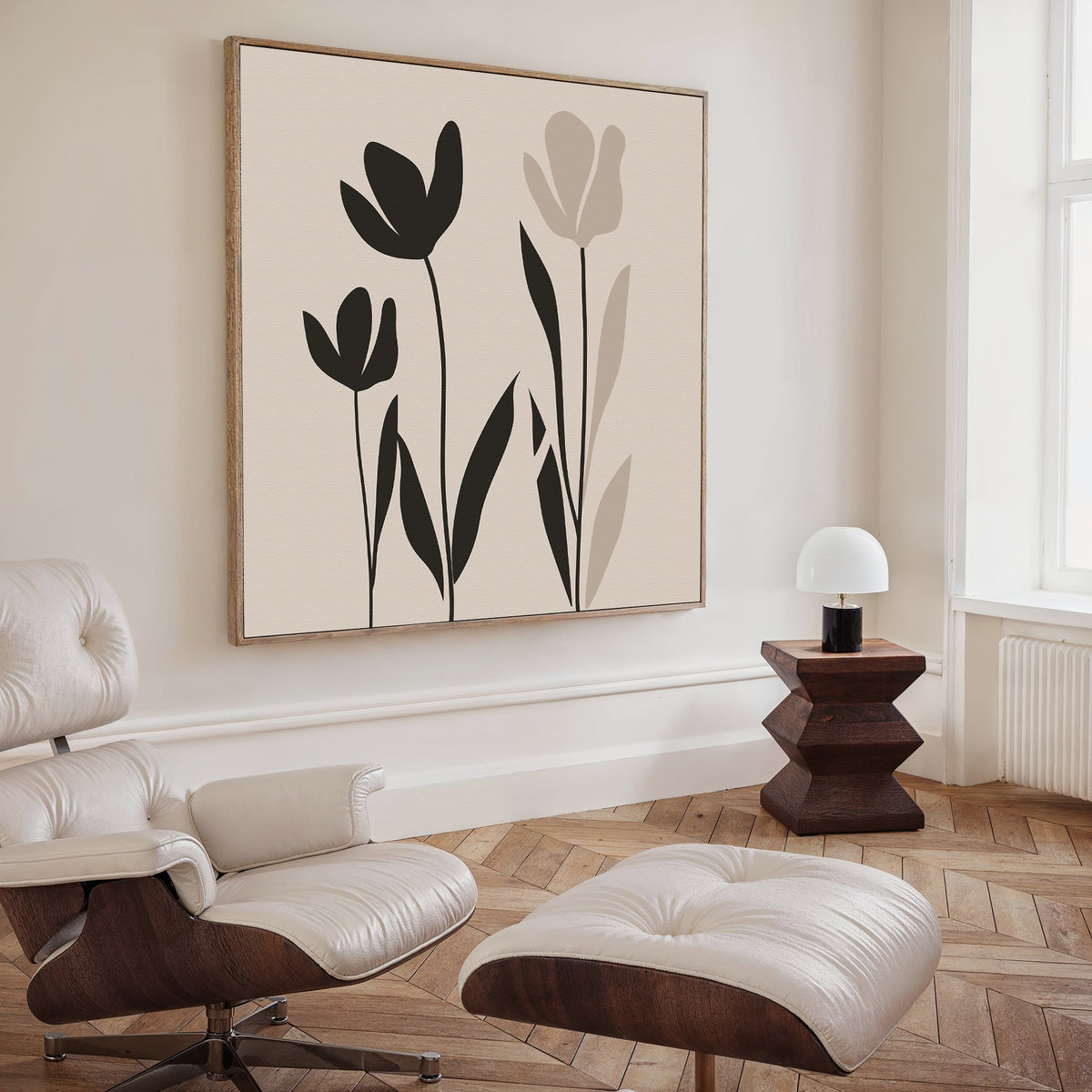 A modern living room with a cream lounge chair and ottoman, wooden side table with a white lamp, and large minimalist Floral Simplicity Canvas Art featuring black and beige florals for a touch of understated elegance.