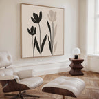 A modern living room with a cream lounge chair and ottoman, wooden side table with a white lamp, and large minimalist Floral Simplicity Canvas Art featuring black and beige florals for a touch of understated elegance.