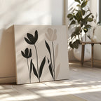 The Floral Simplicity Canvas Art, featuring black and beige tulip silhouettes, leans against a white wall on a sunlit wooden floor, bringing subtle elegance to vintage-inspired decor next to a potted plant and small table.