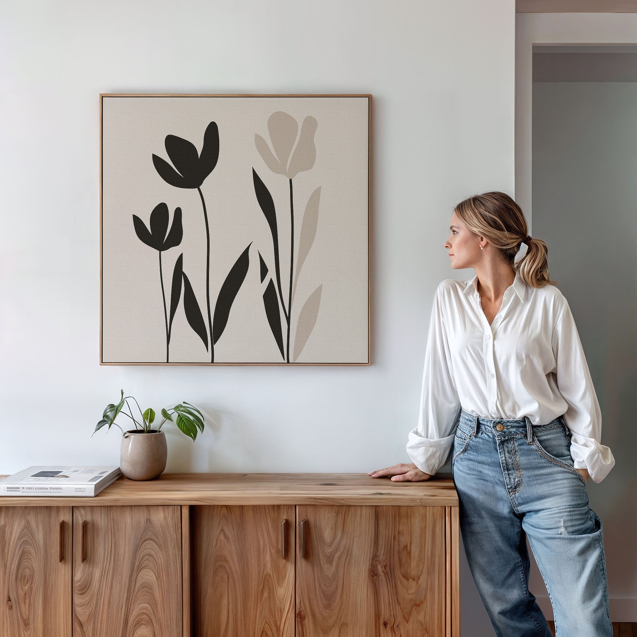 A woman in a white blouse and jeans leans against a wooden cabinet, gazing at the Floral Simplicity Canvas Art above her. A small potted plant and a book rest on the cabinet, enhancing the vintage-inspired decor of the scene.