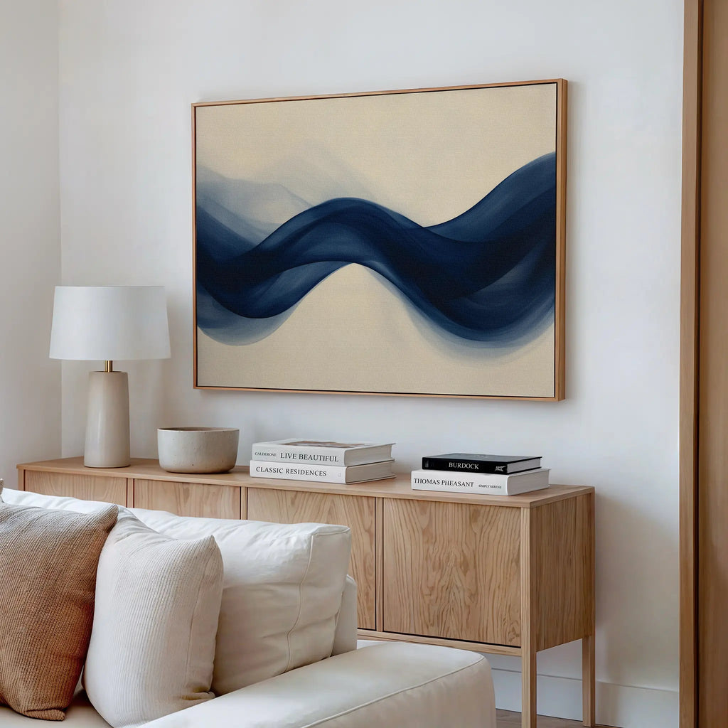 A modern living room features a wooden sideboard with books and decor, a white table lamp, a beige sofa, and the Flowing Indigo Canvas Art—modern wall art of blue waves on a cream background—hanging on the wall.
