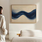 A person in white stands in a minimalist room, gazing at Flowing Indigo Canvas Art—blue waves on beige—hung above a light sofa, bringing a striking touch to the living room décor.