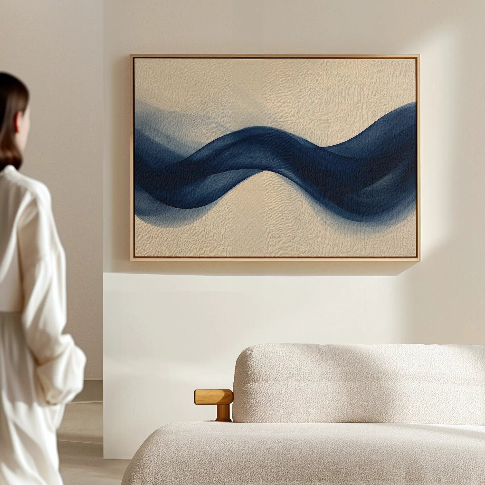 A person in white stands in a minimalist room, gazing at Flowing Indigo Canvas Art—blue waves on beige—hung above a light sofa, bringing a striking touch to the living room décor.