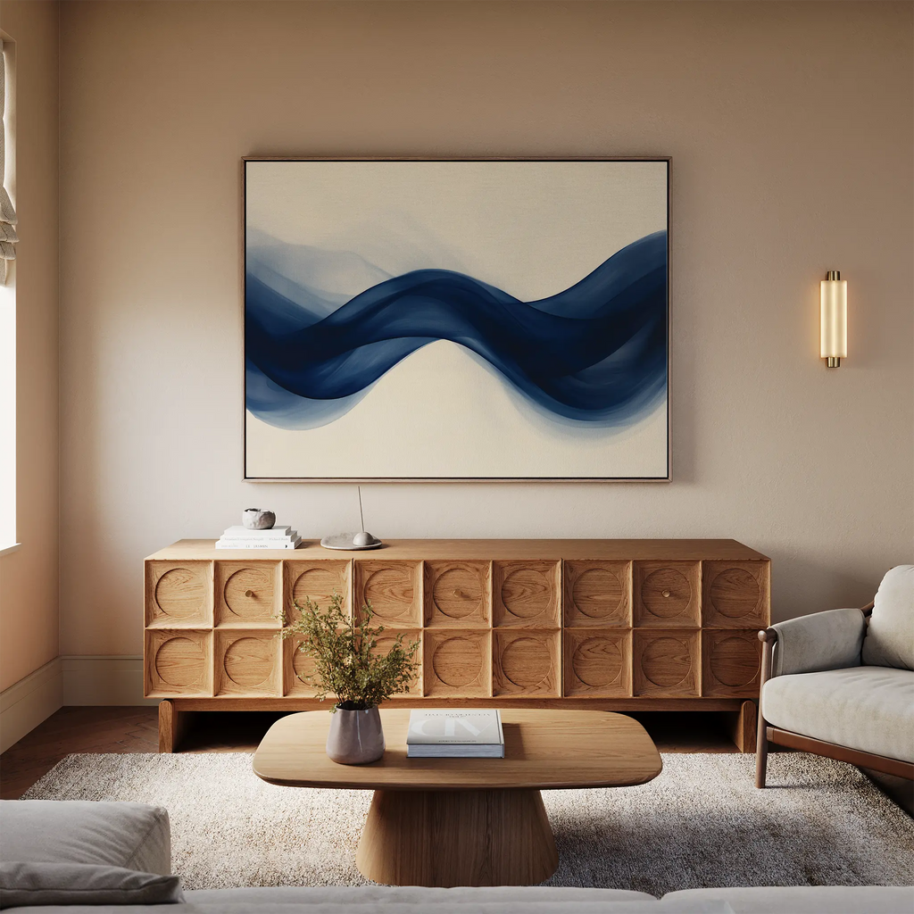 A modern living room with a light sofa, wooden coffee table, decorative credenza, and Flowing Indigo Canvas Art—a striking blue abstract wave painting. A small plant and books complete the stylish minimalist decor.