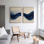 A modern living room with minimalist interiors features a light sofa, woven chair, small round table with a white candle, and the Flowing Indigo Set of 2 wall art. Large windows fill the space with natural light.