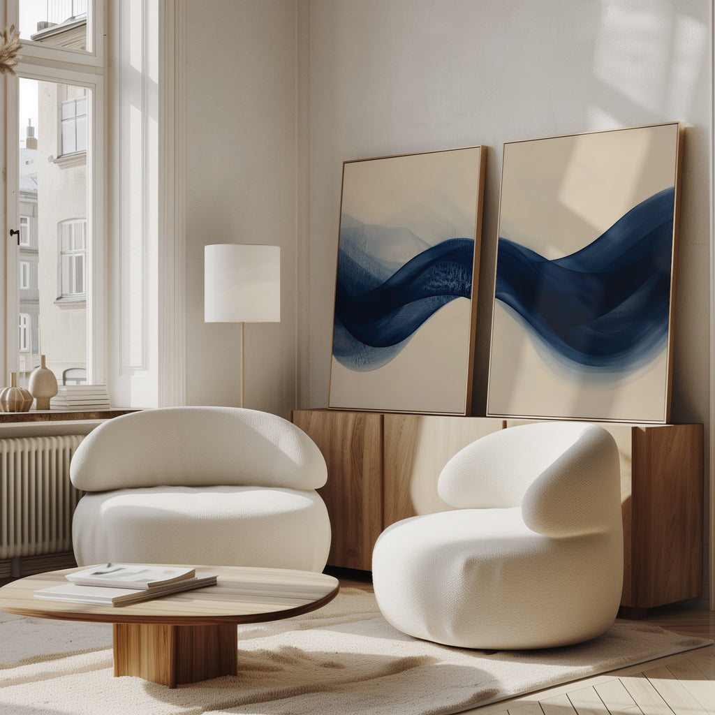 A modern living room with two white rounded armchairs, a low wooden coffee table, and the Flowing Indigo Set of 2 abstract blue wall art leaning against a sideboard. Large windows bring in natural light for a bright, airy feel.