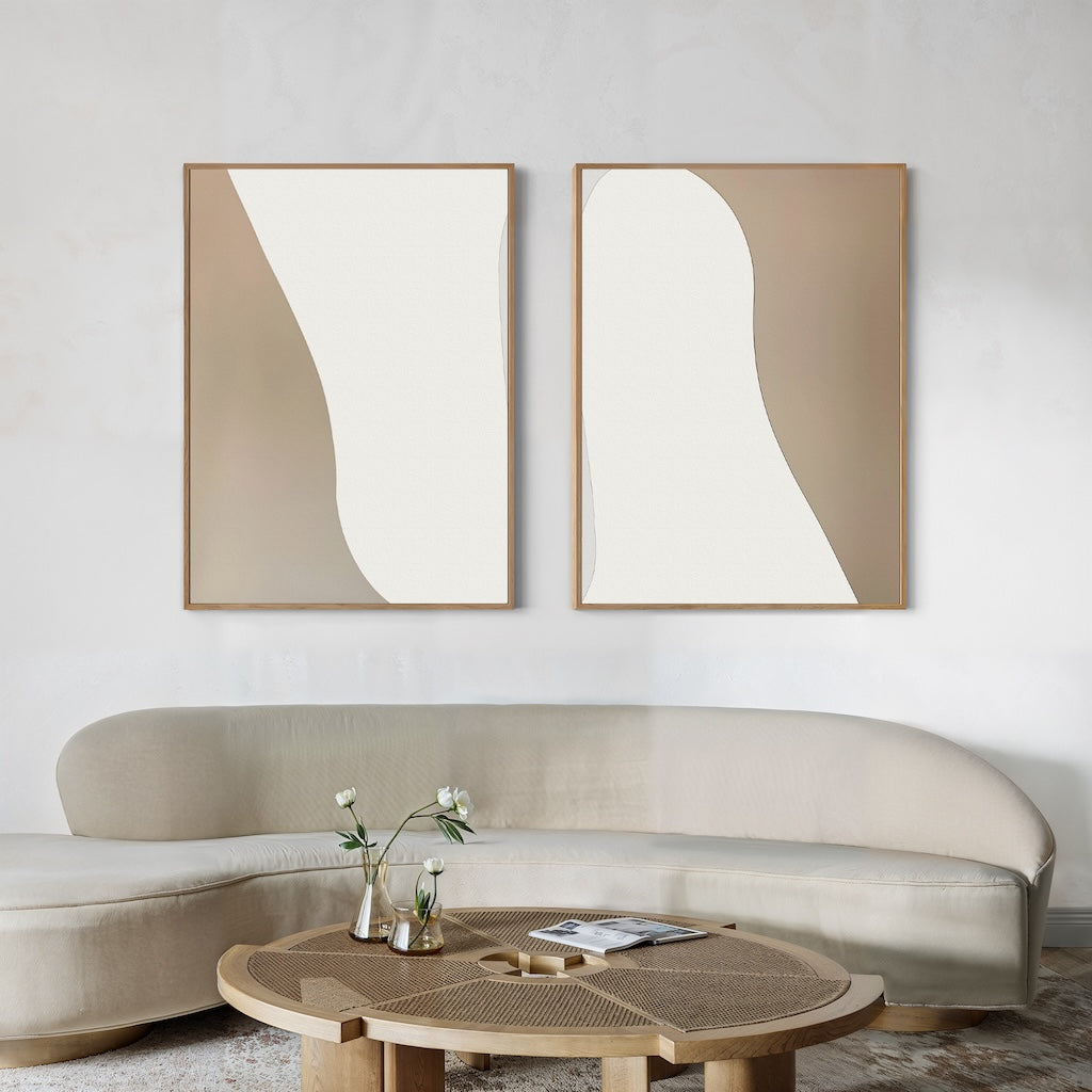 A minimal living room with a curved beige sofa, a round wooden coffee table with vases and books, and the Flowing Silhouettes Set of 2 modern wall art pieces above the sofa—perfect for chic living room decor.