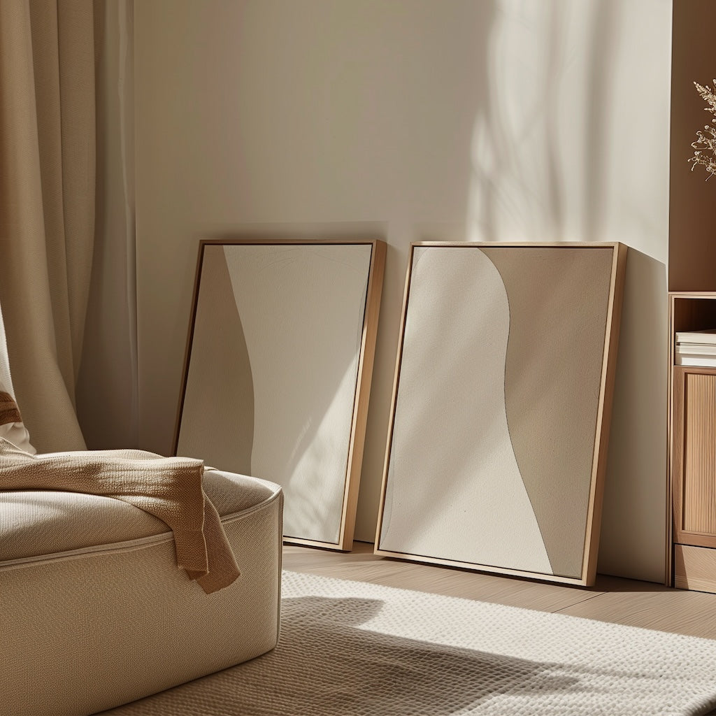 The Flowing Silhouettes Set of 2, featuring minimalist wall art in neutral tones with wooden frames, leans against a beige wall in a sunlit, cozy room, adding warm decor and natural beauty to your living space.