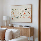 A modern living room features a wooden console table, stacked books, a white lamp, and the Fluttering Dreams Abstract Canvas Art above a beige sofa with white and cream cushions.