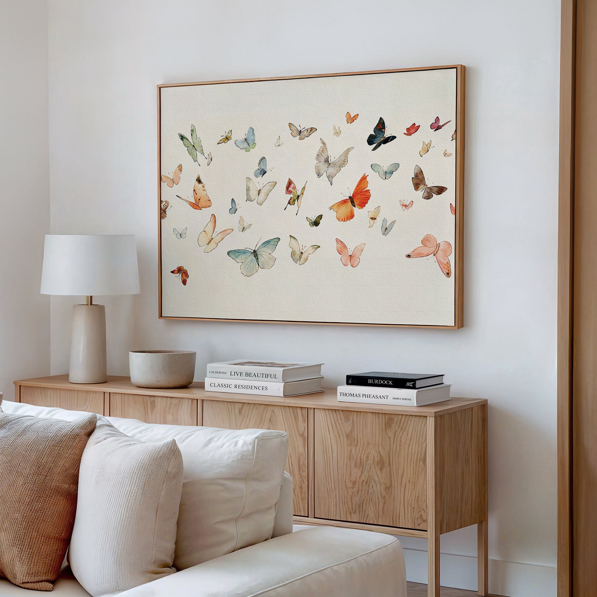 A modern living room features a wooden console table, stacked books, a white lamp, and the Fluttering Dreams Abstract Canvas Art above a beige sofa with white and cream cushions.