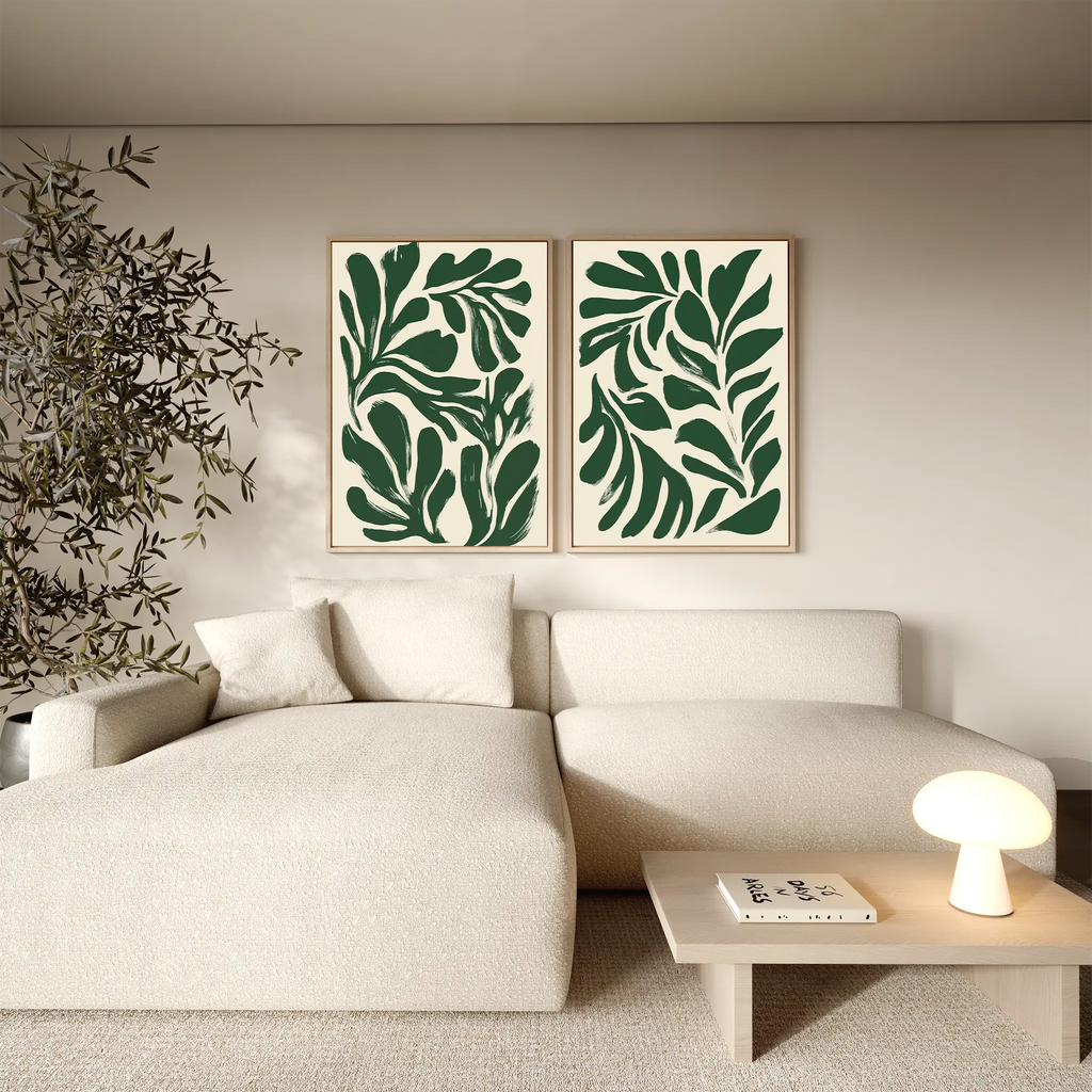 A modern living room with a light beige sofa, a wooden coffee table with a small lamp, a potted plant, and the Forest Leaves Set of 2 framed botanical wall art prints of abstract green leaves hanging above the sofa.