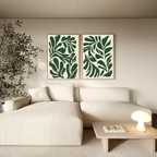 A modern living room with a light beige sofa, a wooden coffee table with a small lamp, a potted plant, and the Forest Leaves Set of 2 framed botanical wall art prints of abstract green leaves hanging above the sofa.