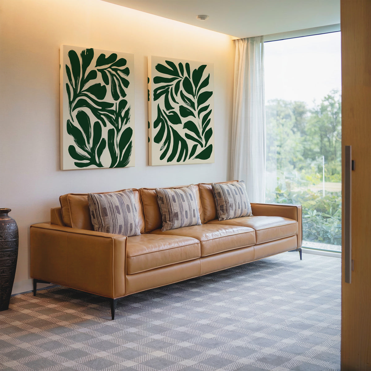 A modern living room with a tan leather sofa, patterned pillows, a Forest Leaves Set of 2 botanical wall art, a large window with sheer curtains, and a decorative vase on the floor.