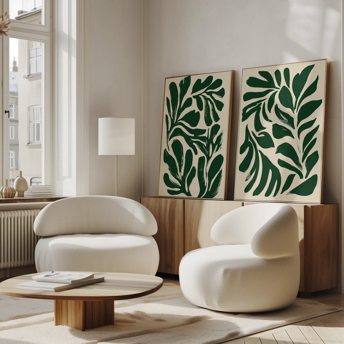 A modern living room with two curved white chairs, a low wooden table, wood sideboard, large windows, and the Forest Leaves Set of 2 botanical wall art. Natural light fills the minimalist space accented by green prints of forest leaves.