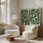 A modern living room with two curved white chairs, a low wooden table, wood sideboard, large windows, and the Forest Leaves Set of 2 botanical wall art. Natural light fills the minimalist space accented by green prints of forest leaves.