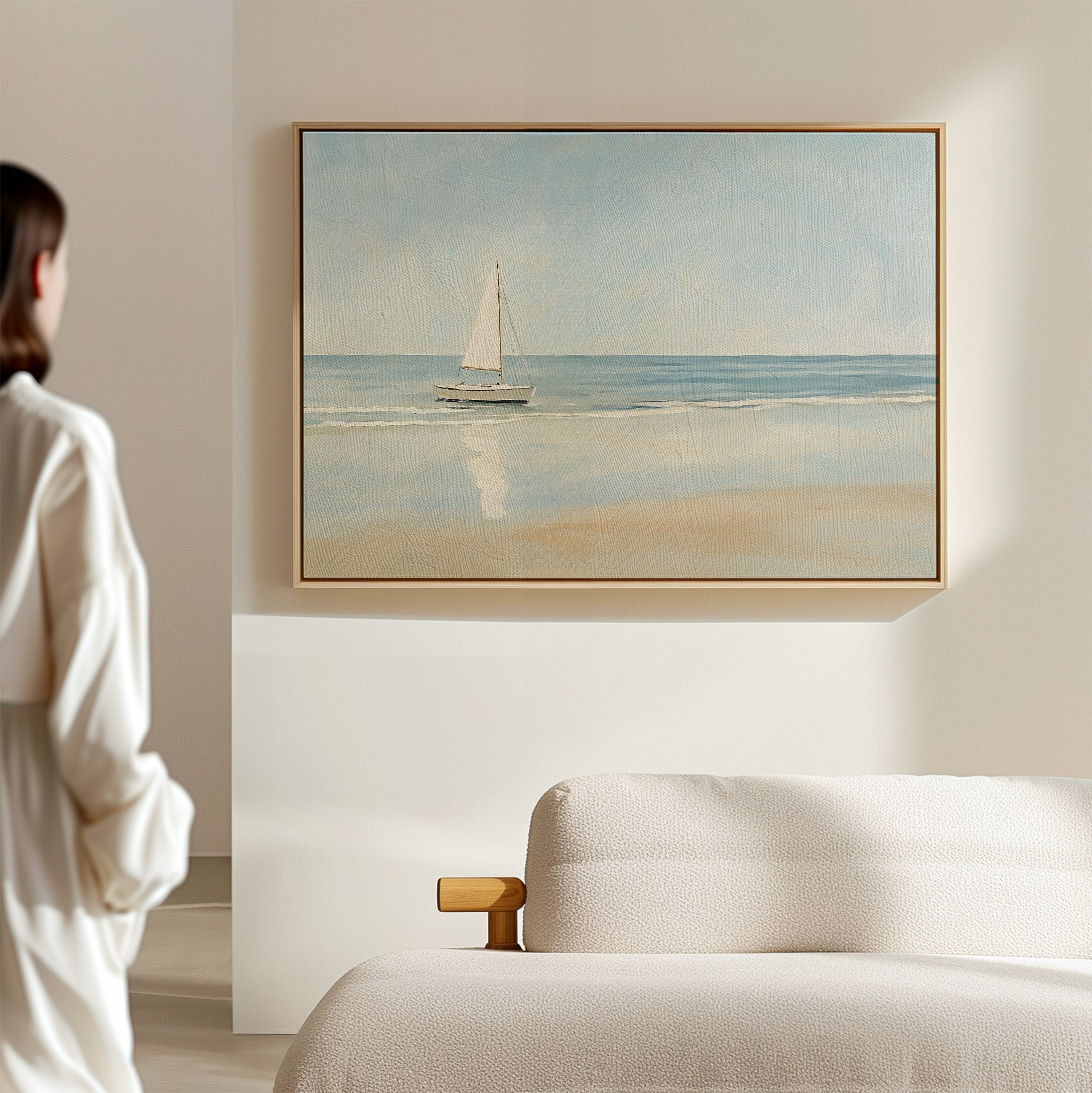 A woman in a white top stands in a sunlit, minimal room, admiring the Freedoms Call Canvas Art—a serene sailboat print above a light-colored sofa that brings tranquil decor to the space.