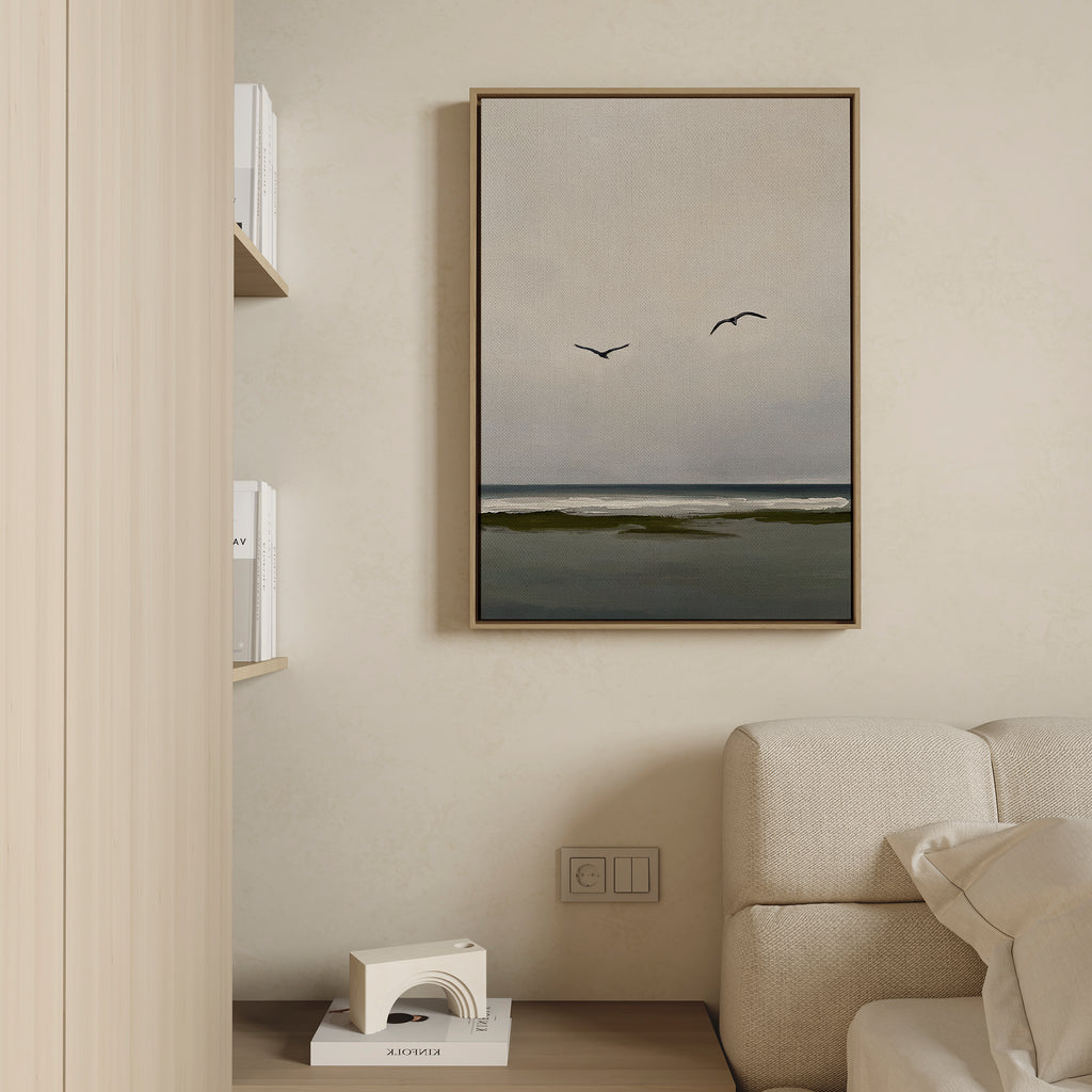 A minimalist living room showcases serene decor, highlighted by the Freeflight Canvas Art of the sea and two birds above a light sofa. Shelves nearby display books, with a small white sculpture on a lower shelf.