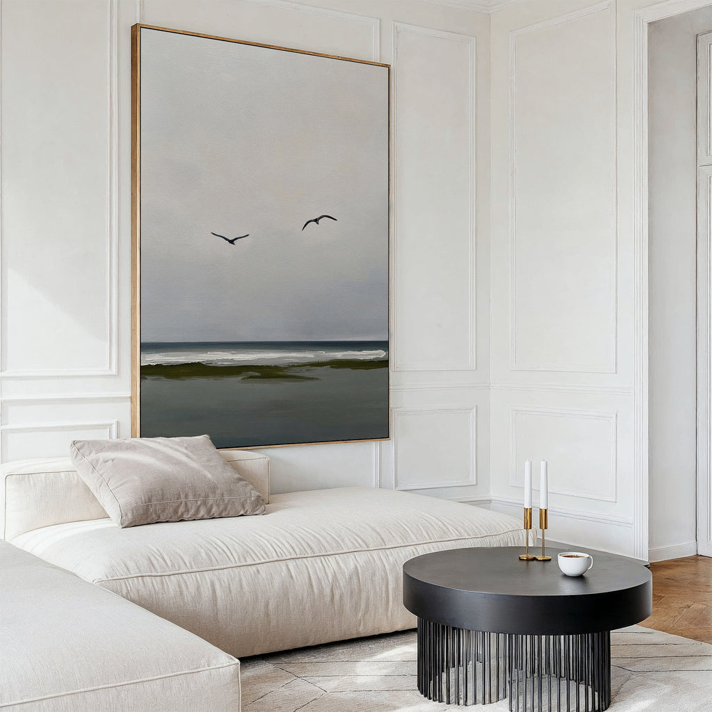 A modern, minimalist living room features a cream sofa, black round coffee table, candles, cup, and serene decor highlighted by the Freeflight Canvas Art—large wall art depicting an ocean view with two birds flying.