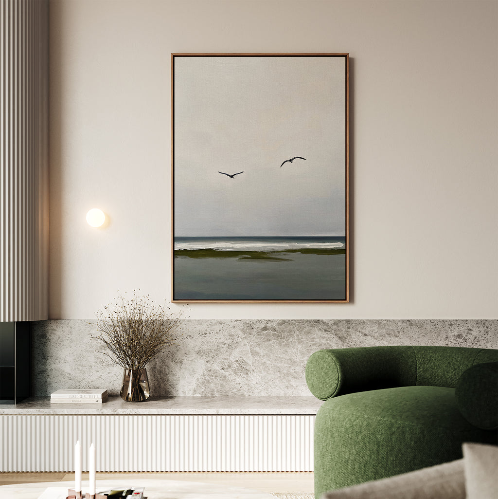 A modern living room features the Freeflight Canvas Art—an ocean painting with two birds—framed and hung above a marble shelf with a vase of dried flowers; a green armchair completes the serene decor.