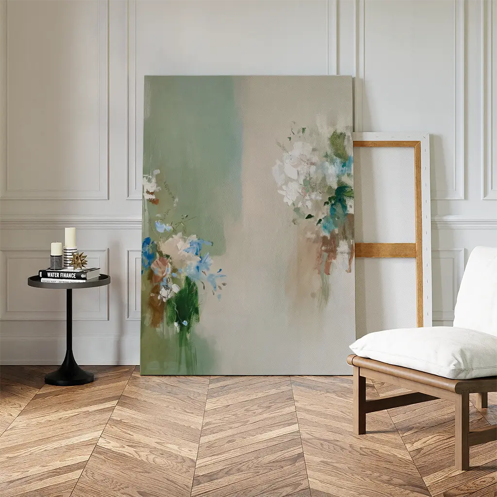 The Garden Bloom Canvas Art leans against a white paneled wall beside an empty frame. A small black round table with candles and books, plus a cushioned chair on the herringbone wood floor, add elegant decor.
