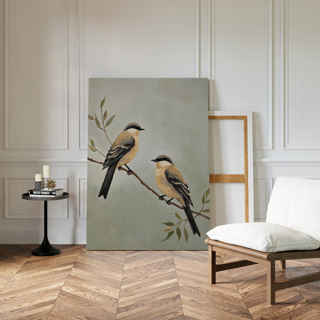 The Garden Pair Canvas Art, featuring two birds on a branch, leans against a white wall in a chic room with wood floors, a cushioned chair, and a small black side table—ideal for Nordic Park-inspired living room decor.