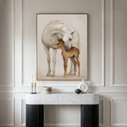 The Gentle Bond Canvas Art, featuring a white horse nuzzling a brown foal in neutral tones, is displayed on an elegant cream-paneled wall above a marble console table adorned with modern decor.