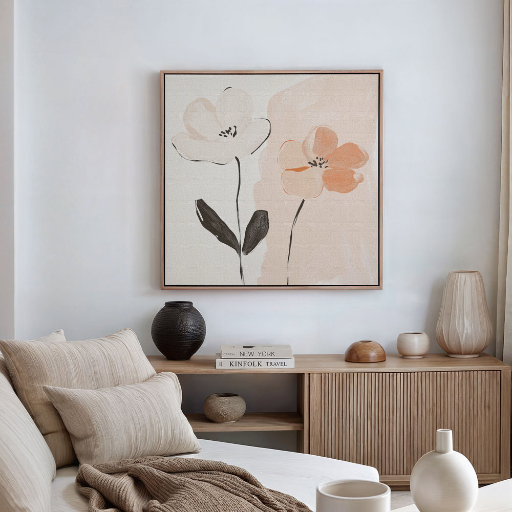 A modern minimalist living room with a beige sofa, neutral pillows, wooden sideboard, pottery, books, and the Gentle Duo Canvas Art—a framed painting of two simple flowers in muted tones—displayed on a white wall.