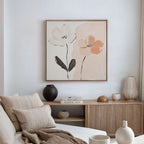 A modern minimalist living room with a beige sofa, neutral pillows, wooden sideboard, pottery, books, and the Gentle Duo Canvas Art—a framed painting of two simple flowers in muted tones—displayed on a white wall.