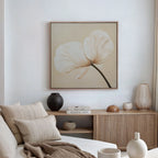 A minimalist living room with neutral tones showcases a beige sofa, a wooden sideboard, and the Gentle Poppy Canvas Art—a framed print of a white two-petal flower—adding an elegant floral touch to the space.