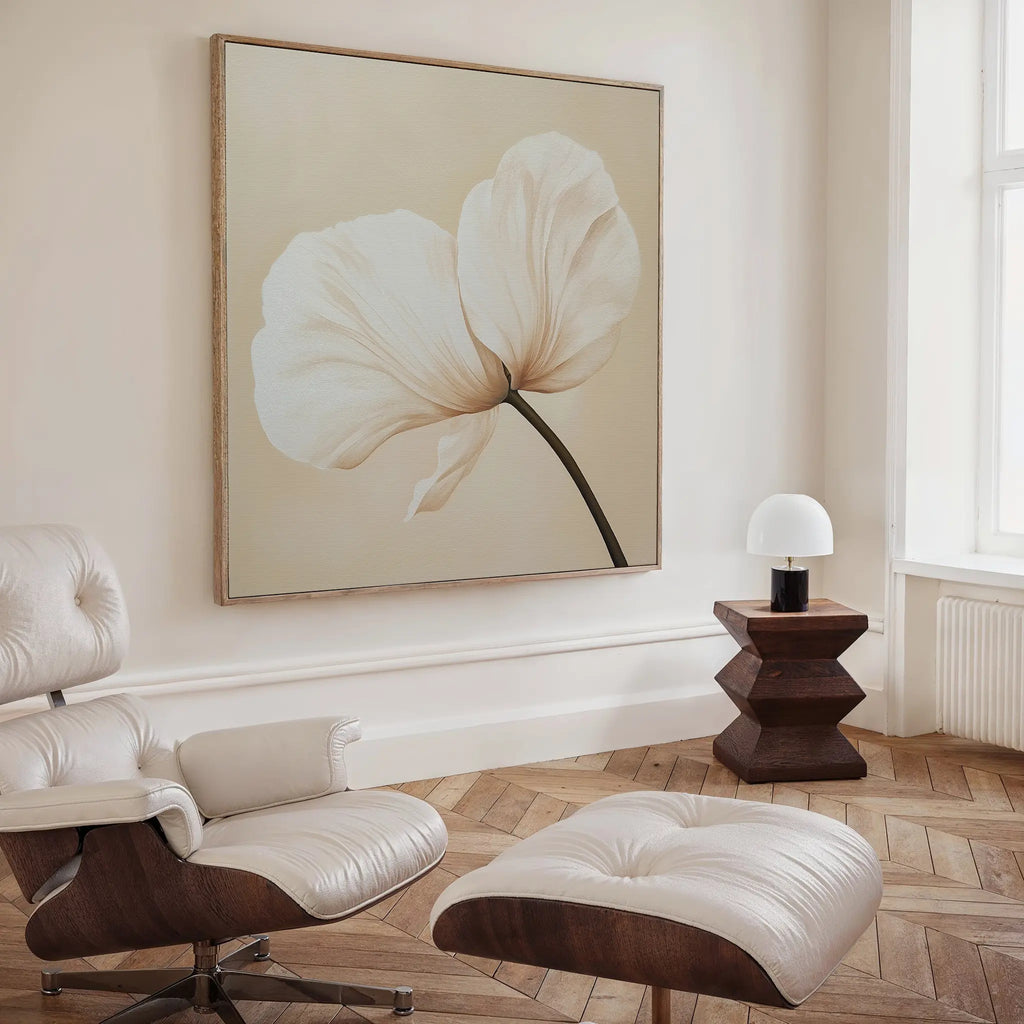 A modern, minimalist room showcases the Gentle Poppy Canvas Art in soft beige tones, complemented by a white leather lounge chair and ottoman, a geometric wooden side table, and a small lamp beside a tall window.