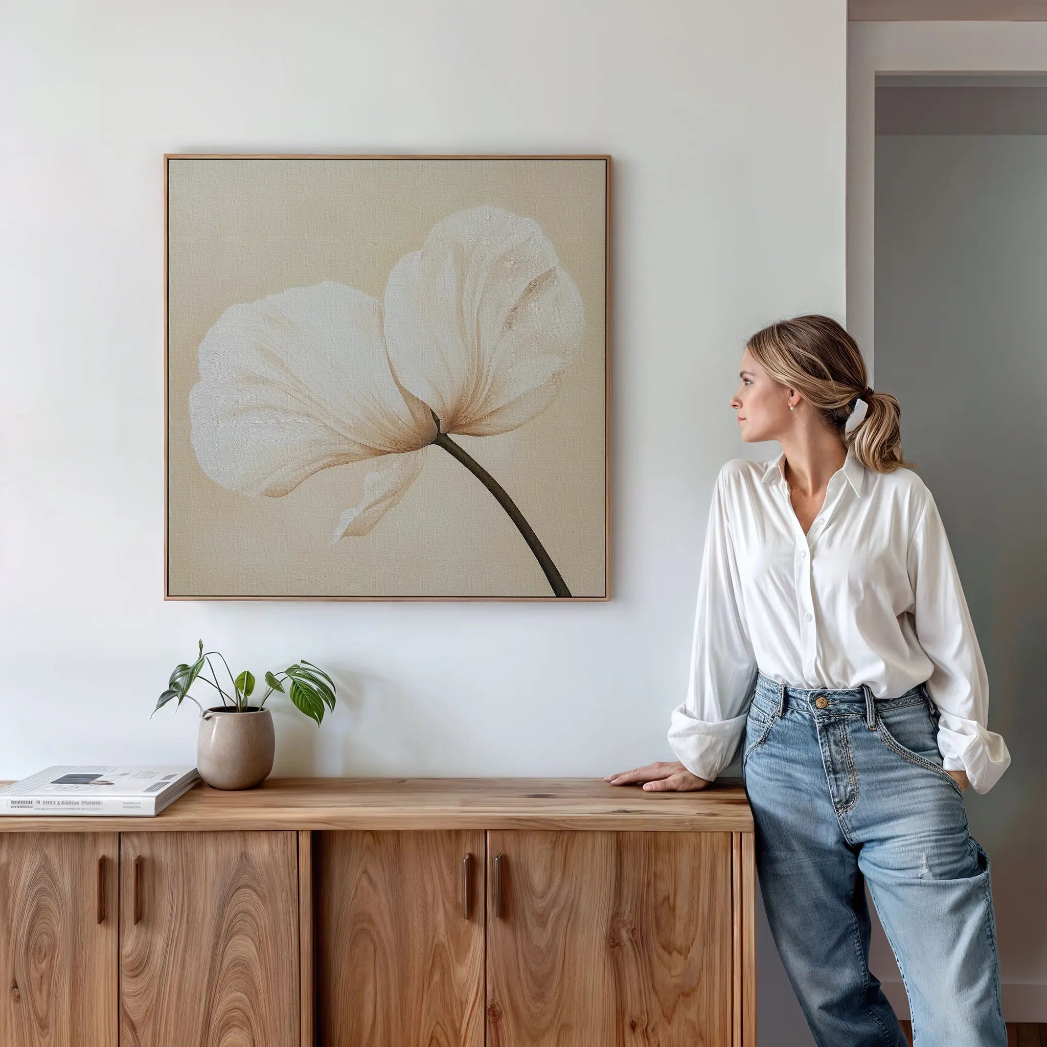 A woman in a white blouse and jeans leans on a wooden cabinet, gazing at the Gentle Poppy Canvas Art above. A small potted plant and a book sit on the cabinet, enhancing the room’s minimalist decor.