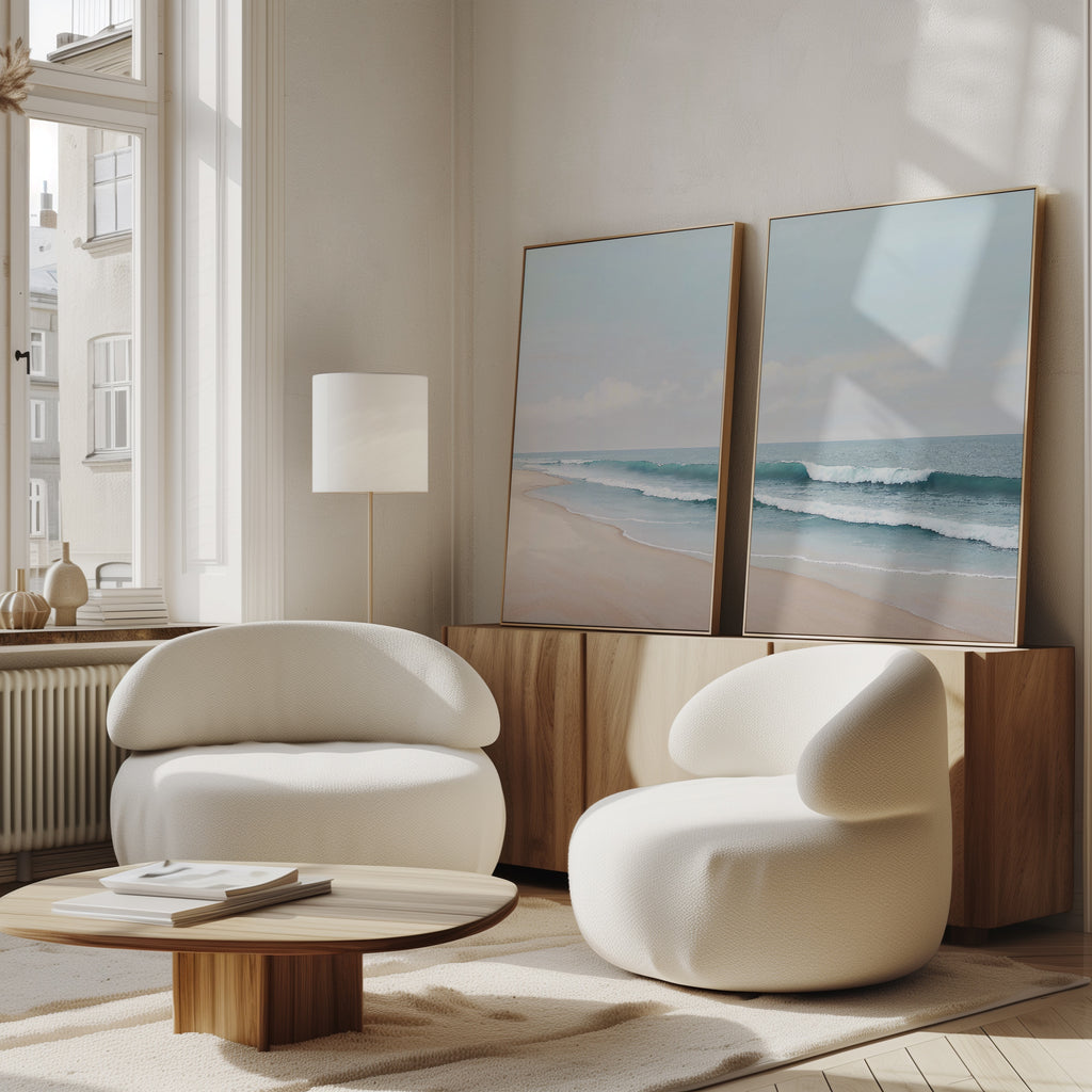 A modern living room featuring the Gentle Surf Set of 2 coastal wall art on a wooden sideboard, with two white rounded armchairs, a low wooden coffee table, and sunlight streaming through tall windows for a bright, airy atmosphere.