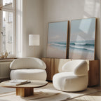 A modern living room featuring the Gentle Surf Set of 2 coastal wall art on a wooden sideboard, with two white rounded armchairs, a low wooden coffee table, and sunlight streaming through tall windows for a bright, airy atmosphere.