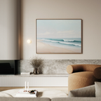 A modern living room featuring a beige curved armchair, marble shelves, and Gentle Surf Canvas Art—a large framed beach scene—hanging on a light wall above a vase of dried flowers; perfect for stylish decor.