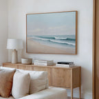 A serene, minimalist living room features a light wood sideboard, white table lamp, decorative bowls, stacked books, and the Gentle Surf Canvas Art—a large framed beach and ocean painting—above a white sofa for perfect decor.