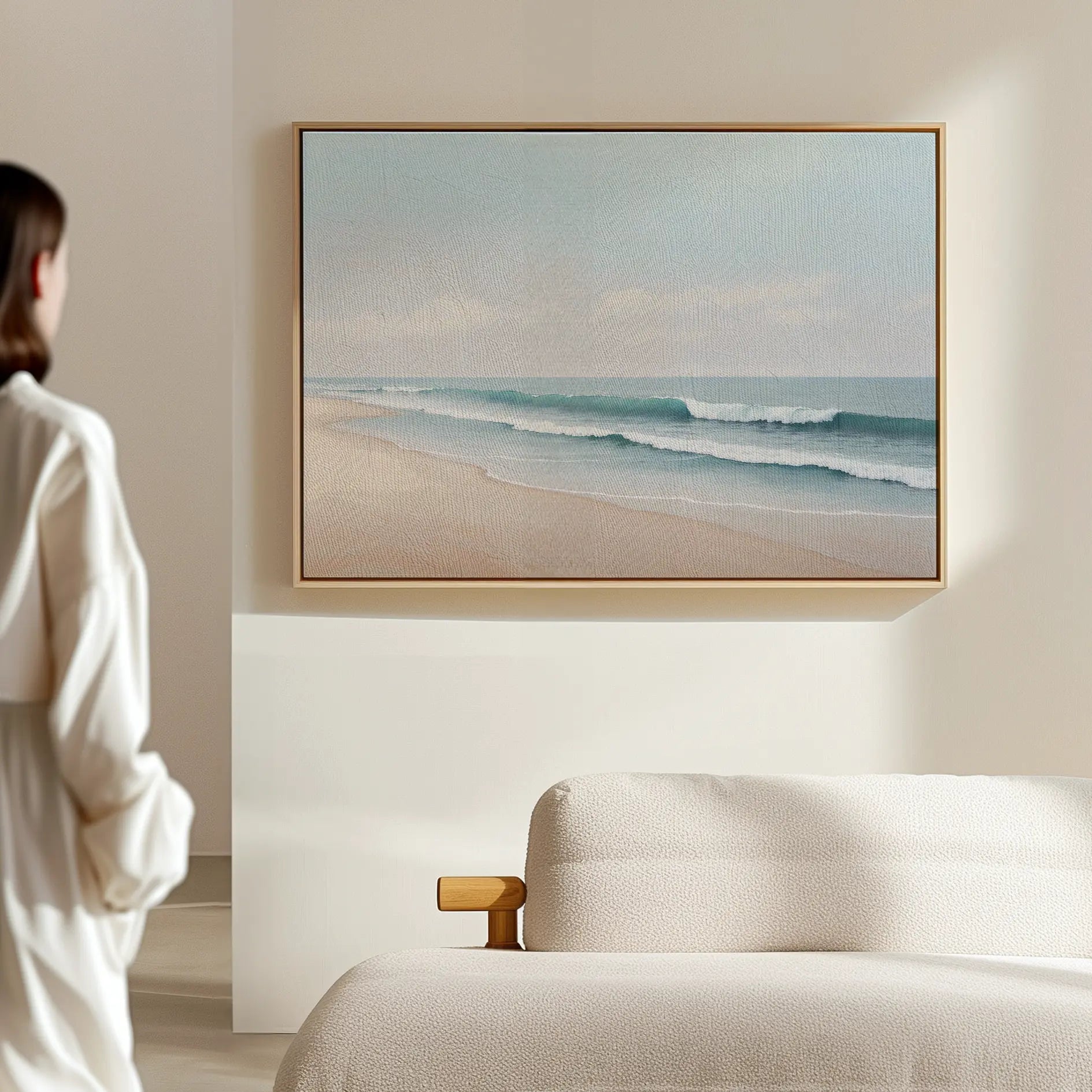 In a minimal, neutral-toned room, a person in white admires the Gentle Surf Canvas Art—framed waves and pale sky—hung above a light sofa, creating serene living room decor.